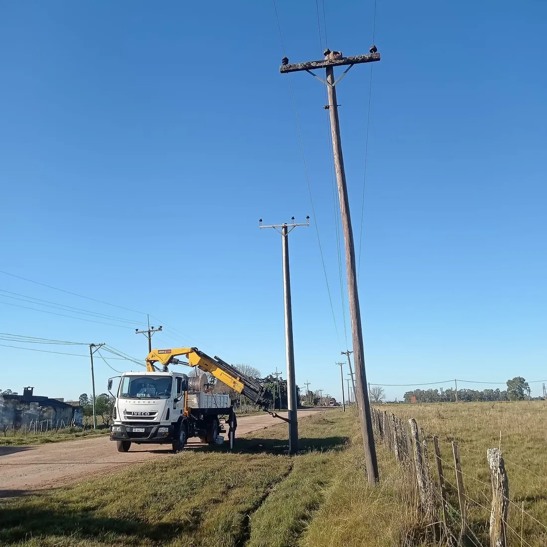 Un poste de madera inclinado junto a la columna de hormigón ya plantada, con el camión con hidrogrúa de la Cooperativa detenido en el camino rural